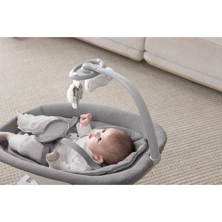 Baby in a gray baby bouncer with a mobile on a beige carpet